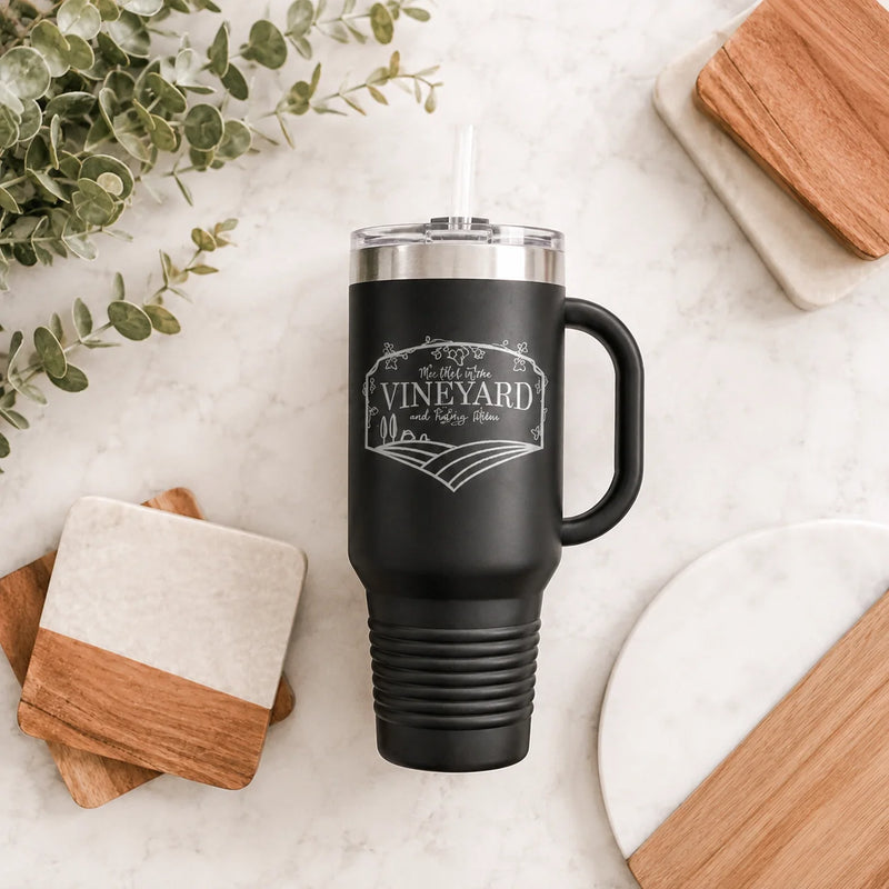A matte black insulated tumbler with a handle, clear lid, and straw, printed with a white vineyard-themed graphic reading 'The Best VINEYARD and Winery Home.' It is photographed in a styled flat lay on a light marble surface with wood and stone coasters, a plate, and greenery around it.