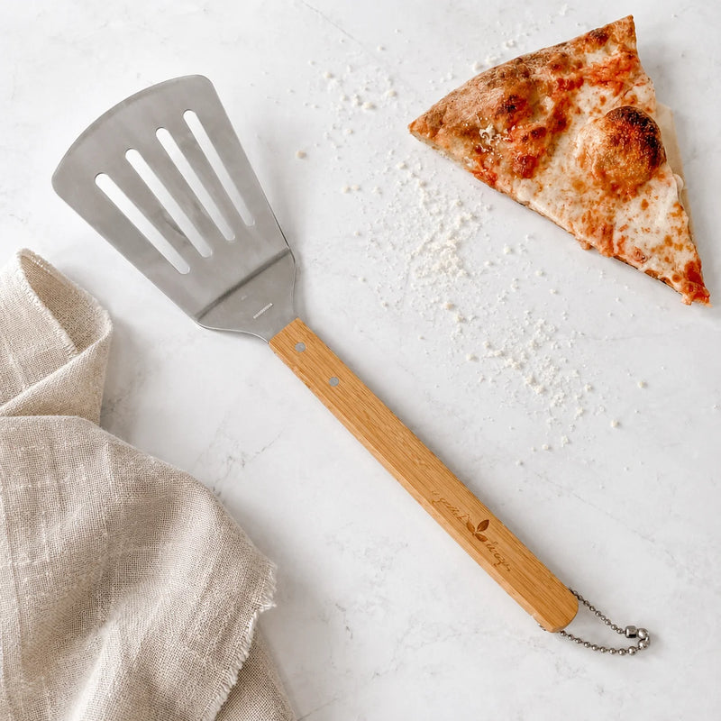 A metal pizza spatula with a light wood handle lies on a white marble surface next to a slice of pizza and a beige linen cloth. The spatula has a silver metal head with slots and a natural wood handle.