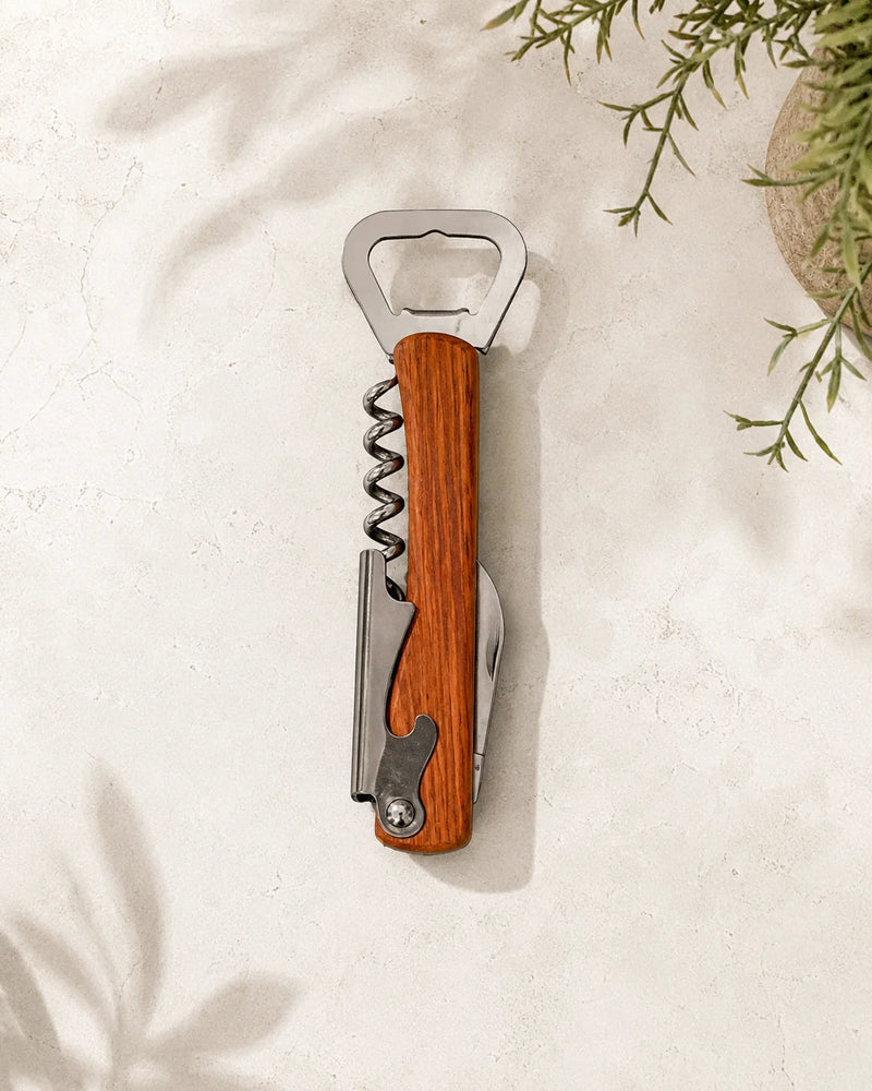 A multi-tool bottle opener with a warm brown wood handle and silver stainless steel blades, shown in a flat-lay style on a textured off-white surface with soft plant shadows.