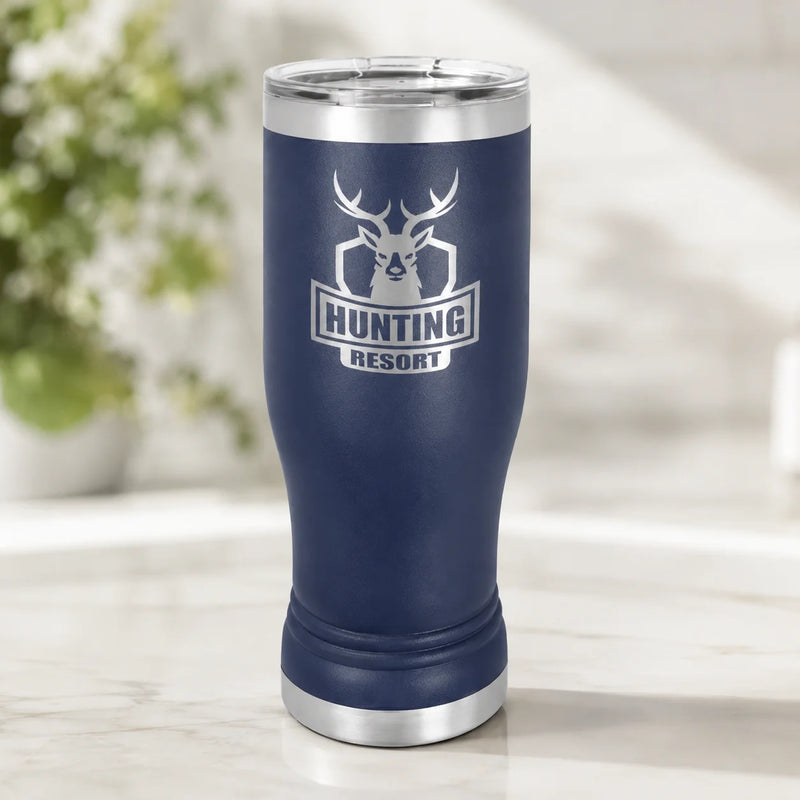 A navy blue insulated travel tumbler with a stainless steel lid and base, featuring a white deer head logo and the text 'HUNTING RESORT' on the front, set on a light marble surface with a blurred indoor background.