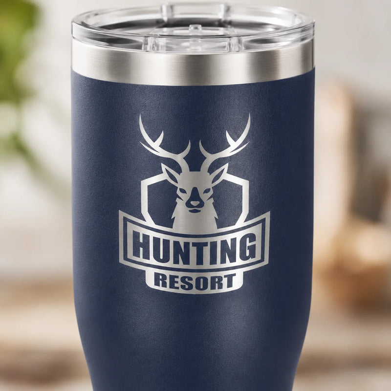 A navy blue insulated tumbler with a clear lid, featuring a white deer head logo and the words 'HUNTING RESORT' on the front, shown against a softly blurred outdoor background.