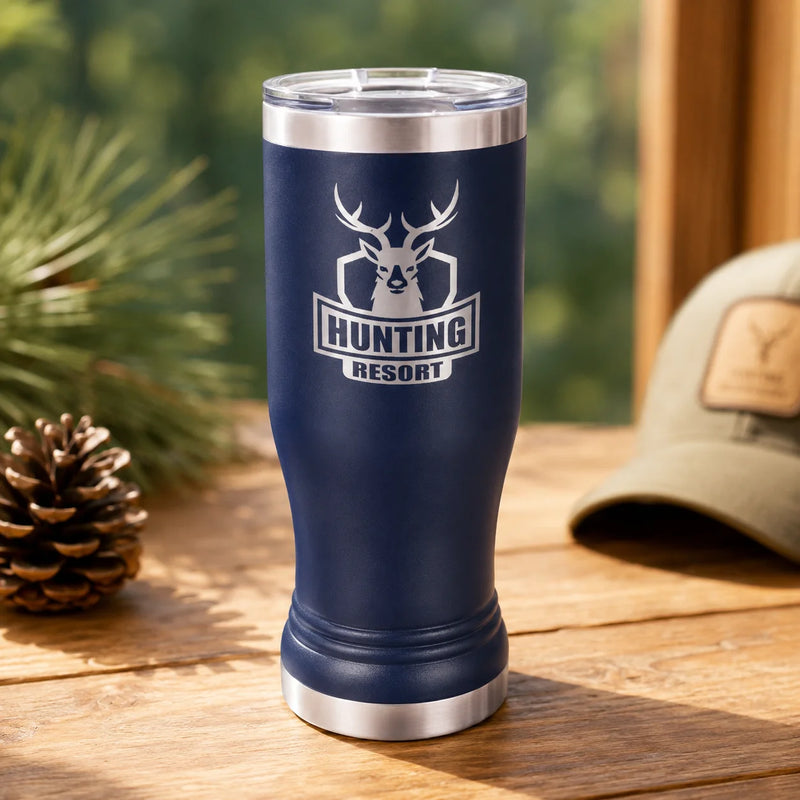 A navy blue insulated tumbler with a clear lid stands on a wooden table. The front has a deer head logo above the words “HUNTING RESORT” in white.