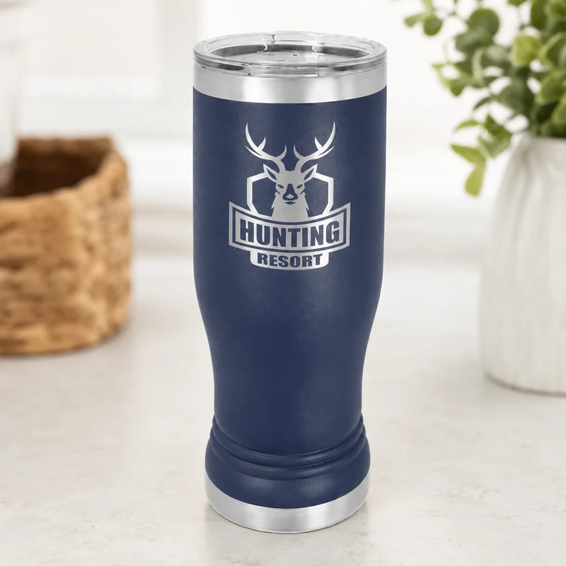 A navy blue insulated tumbler with a silver stainless steel lid and base, featuring a deer head logo and the words 'HUNTING RESORT' on the front, set on a light countertop with blurred background decor.