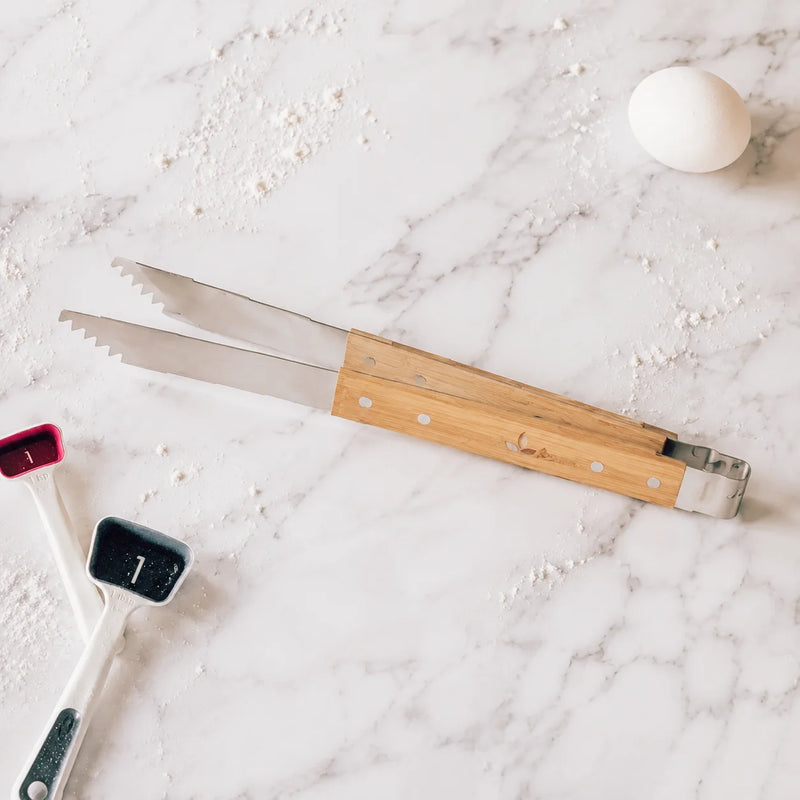 A pair of stainless steel kitchen tongs with a light bamboo handle, laid diagonally on a white marble surface scattered with flour, next to measuring cups and a white egg.