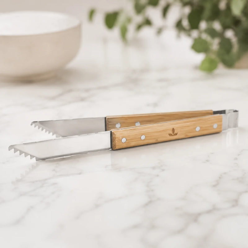 A pair of stainless steel tongs with serrated edges and light natural bamboo handles, resting on a white marble countertop with a blurred kitchen background.