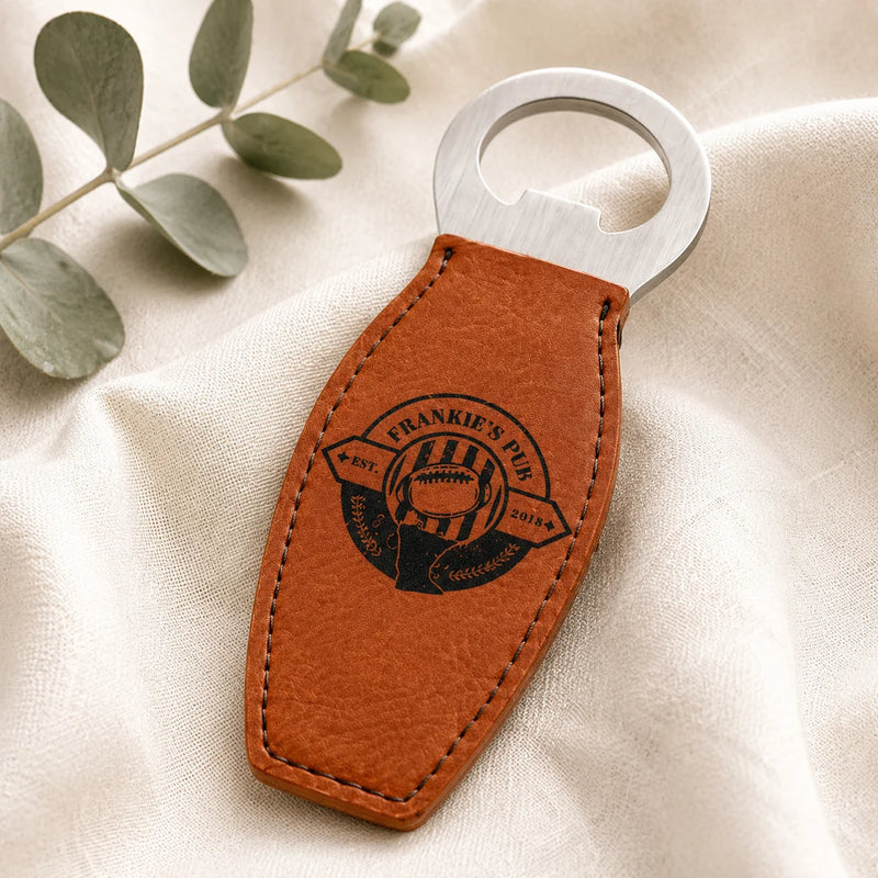 A personalized brown leather bottle opener with a brushed silver metal loop, featuring an engraved football-themed emblem that reads 'FRANKIE'S PIS' and '2024'. The opener is styled on a cream-colored textured fabric with a green eucalyptus sprig in the background.