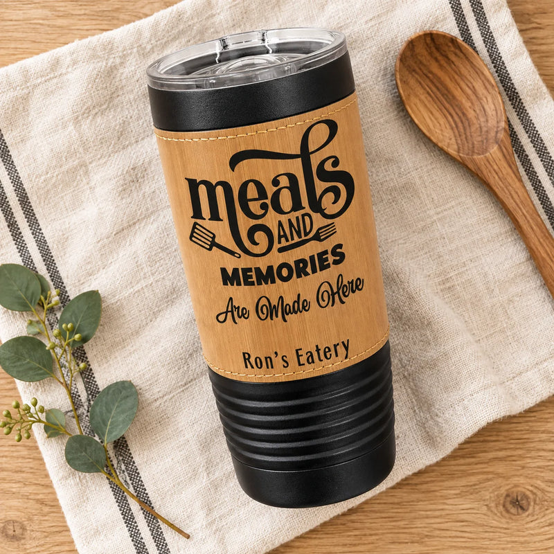 A personalized stainless steel tumbler with a bamboo-style wrap, shown on a striped linen napkin with a wooden spoon and eucalyptus sprig. The tumbler features the engraved text 'meals AND MEMORIES Are Made Here Ron's Eatery'.