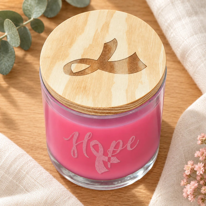 A pink candle in a clear glass jar with a light wood lid engraved with a ribbon, placed on a wooden surface with beige fabric and small flowers around it. The jar is etched with the word 'Hope' and a ribbon motif.