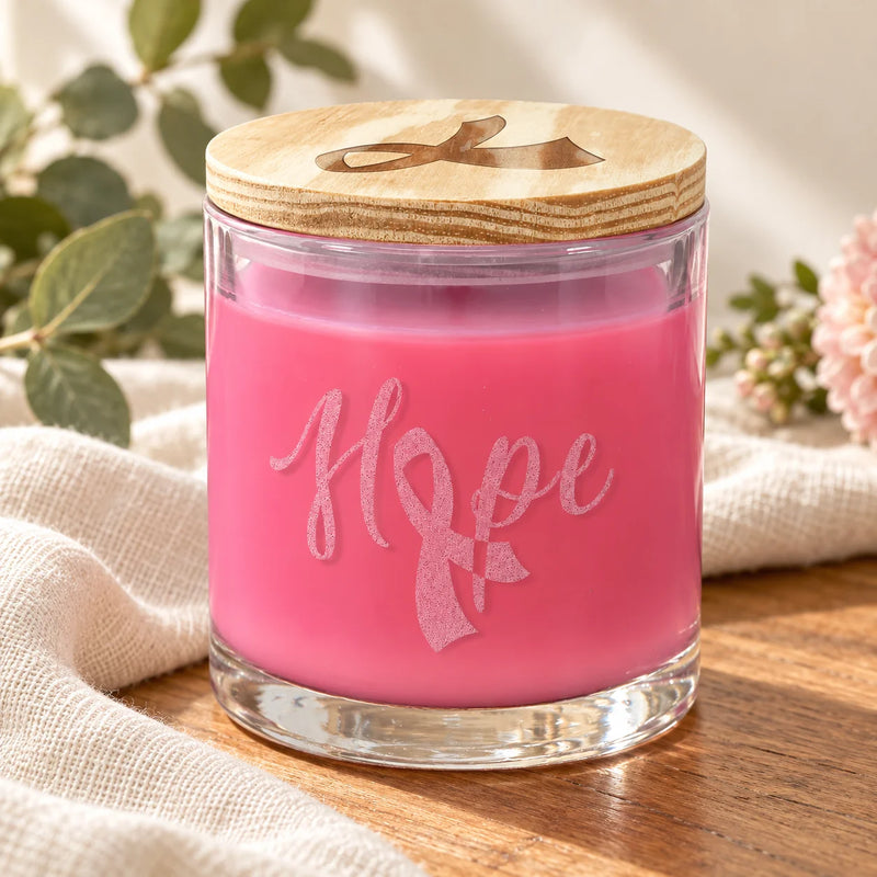 A pink candle in a clear glass jar with a light wood lid engraved with a ribbon, set on a wooden surface with a beige cloth and soft flowers in the background. The word 'Hope' is printed on the front.