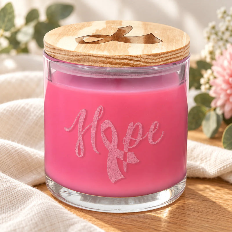 A pink candle in a clear glass jar with a wooden lid and a ribbon-shaped engraving that spells 'Hope' across the front. The candle is styled on a beige cloth over a wooden surface with soft-focus flowers and greenery in the background.