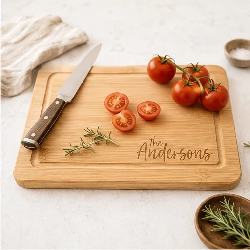 A rectangular bamboo cutting board with a custom engraved name, 'The Andersons', styled on a light marble countertop with a chef's knife, cherry tomatoes, rosemary, and a cloth napkin.