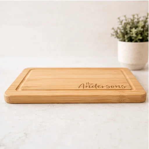 A rectangular bamboo cutting board with a recessed center and the personalized name 'the Andersons' engraved on the front, set on a light marble countertop with a blurred potted plant in the background.