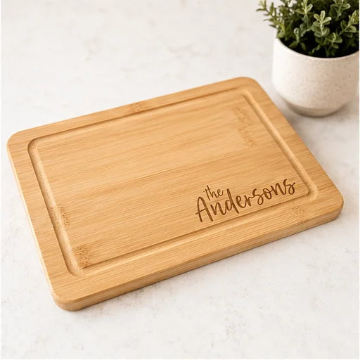 A rectangular bamboo cutting board with rounded corners and a recessed center, engraved with the name 'the Andersons' in script. The board is photographed on a light marble-like surface with a small potted green plant in the background.