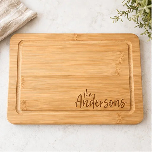A rectangular bamboo cutting board with rounded corners and engraved text reading 'The Andersons' in the lower right, photographed on a light marble surface with a folded cloth and greenery as props.