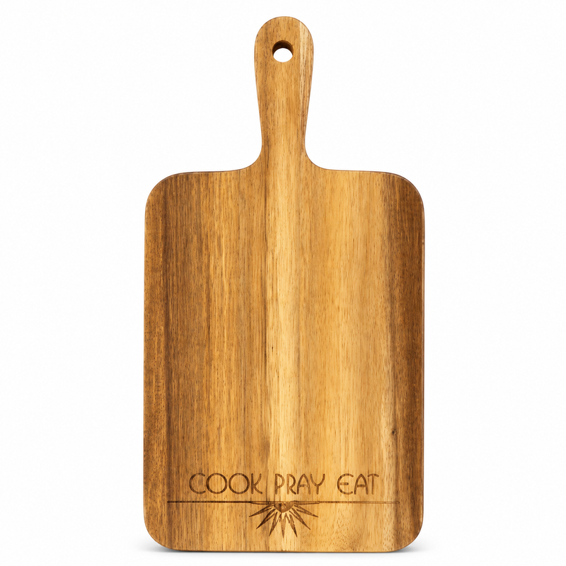 A rectangular wooden cutting board with a handle and a hanging hole, featuring the engraved text 'COOK PRAY EAT' and a small sunburst design near the bottom, shown on a plain white background.