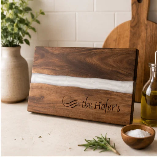 A rectangular wooden cutting board with a white resin inlay and engraved text reading 'the Hofer's', styled on a countertop with a plant, cutting board, oil bottle, and small bowls in a kitchen setting.