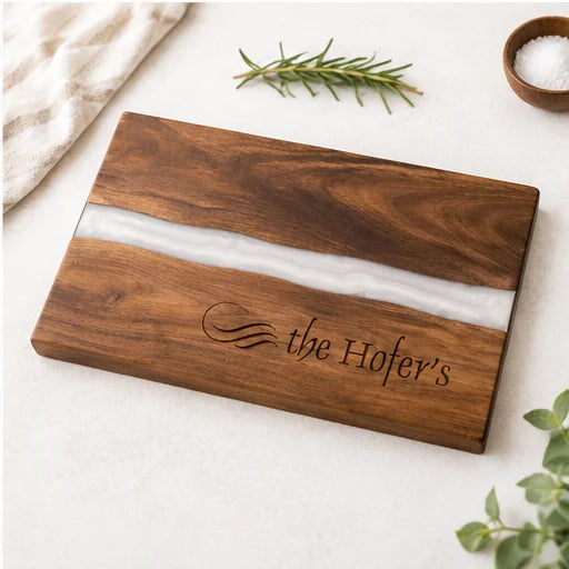 A rectangular wooden cutting board with a white resin inlay and engraved text reading 'the Hofer's', styled on a light surface with a cloth, rosemary, salt, and greenery.
