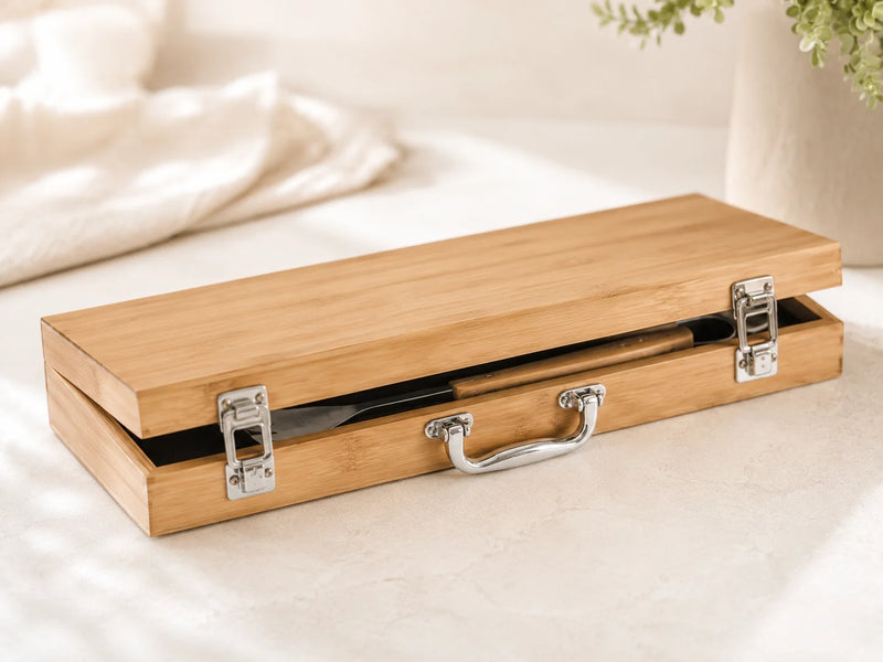 A rectangular wooden presentation box with a hinged lid, silver metal latches, and a curved handle, shown open on a light surface with a softly blurred indoor background.