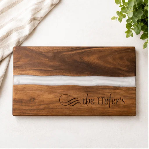 A rectangular wooden serving board with a horizontal white resin inlay and engraved text reading 'the Hofer's', photographed on a light surface with a folded striped cloth and green leaves as props.