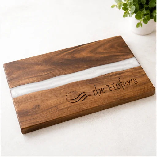 A rectangular wooden serving board with a polished white resin inlay and engraved text reading 'the Hofer's', photographed on a light neutral surface with a small plant in the background.