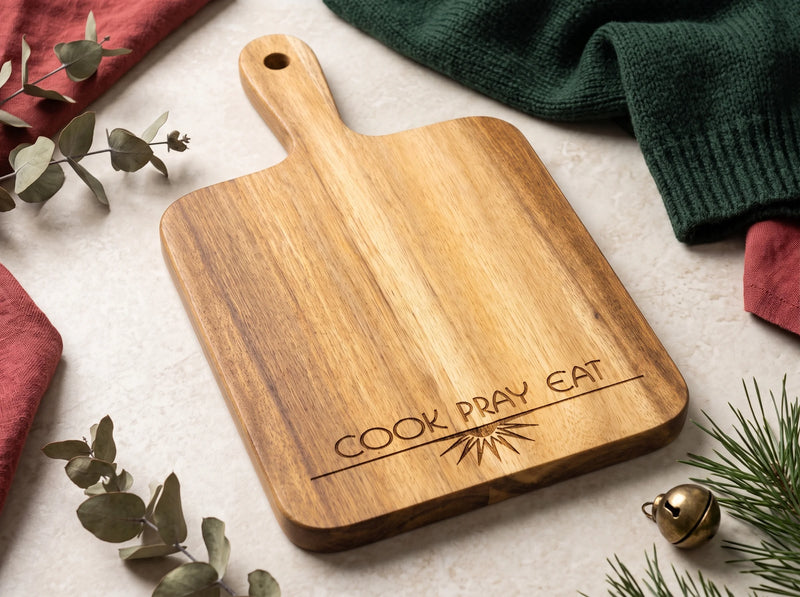 A rectangular wooden serving board with rounded corners and a handle, engraved with the words 'COOK PRAY EAT' and a small sunburst design. The board is styled in a flat-lay setting on a light textured surface with eucalyptus branches, a green knit sweater, red fabric, and a small gold bell.