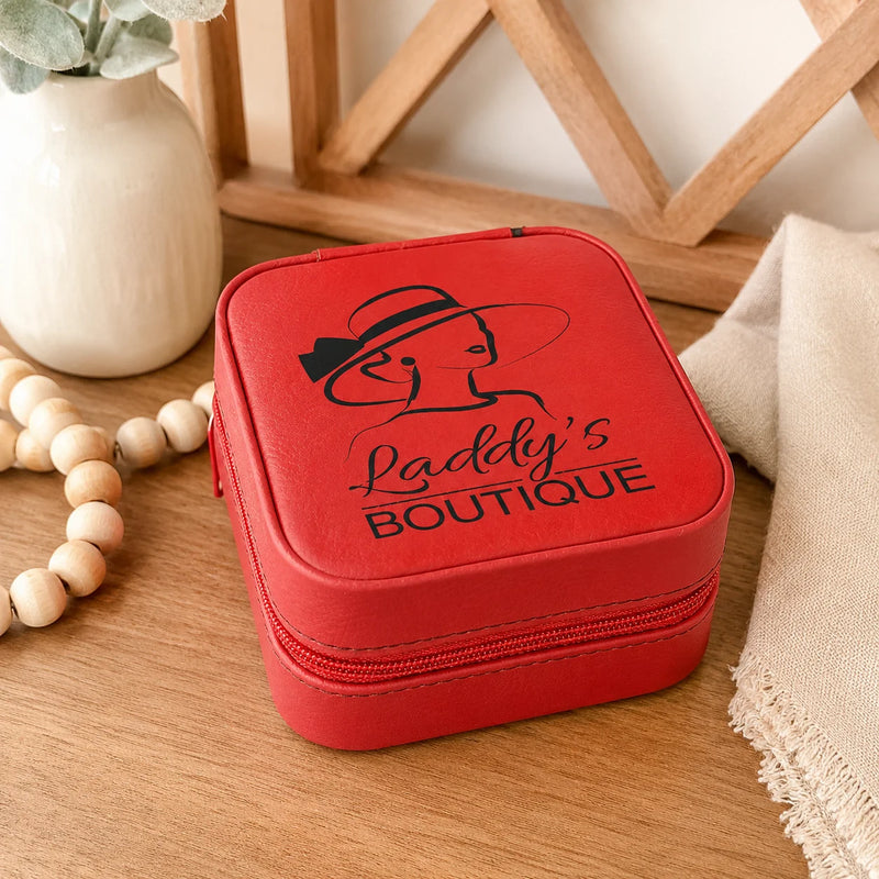 A red faux-leather zippered jewelry case with a black printed logo reading 'Laddy's Boutique' and a hat-wearing woman illustration, placed on a wooden table with decorative props including a vase, beads, and a cloth.