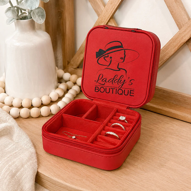 A red zippered jewelry organizer case shown open on a wooden surface, with ring slots, compartments, and a printed 'Lady's Boutique' logo on the inside lid. Decorative beads, a white vase, and a wooden frame are in the background.