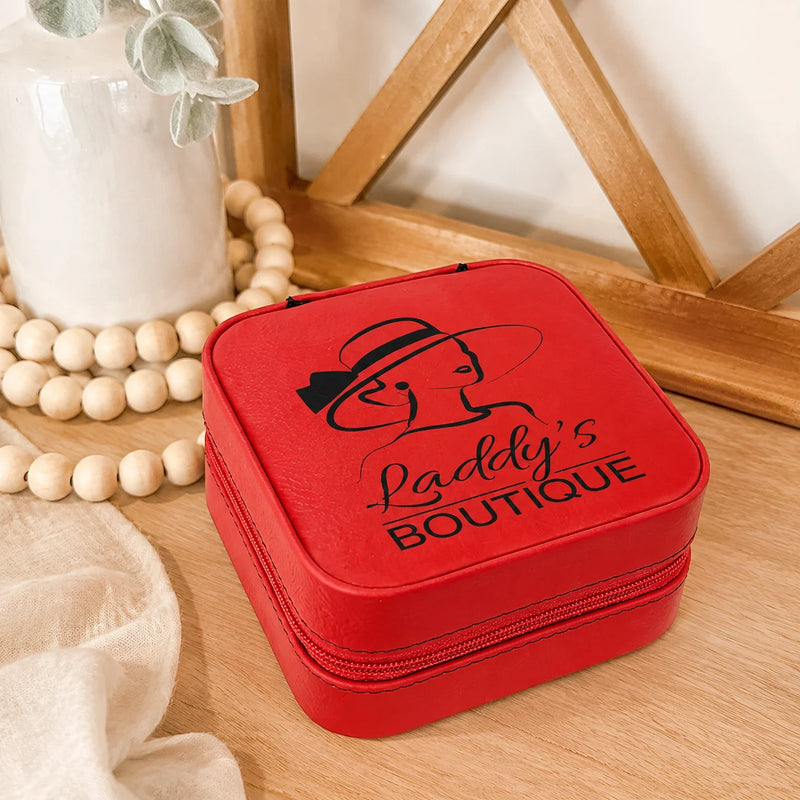 A red zippered jewelry organizer case with a black printed logo and the words 'Laddy's Boutique' on the lid, placed on a wooden surface with decorative beads, a white vase, and a wooden frame in the background.