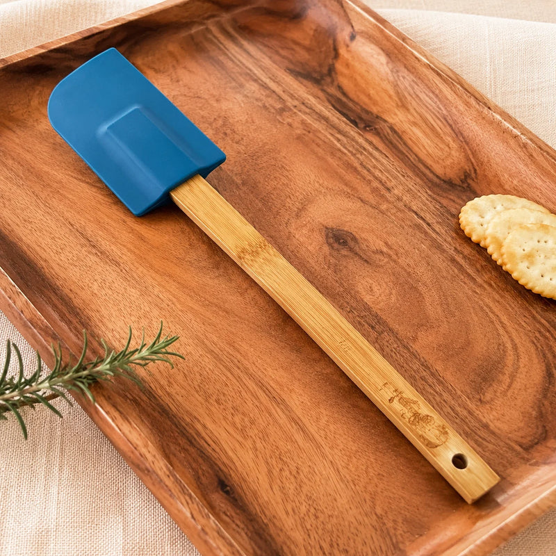 A silicone spatula with a blue head and a light bamboo handle rests diagonally on a rectangular wooden serving tray. The tray is styled with a piece of rosemary and a cracker on a beige fabric surface.