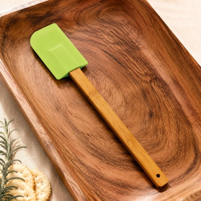 A silicone spatula with a bright green blade and a light brown bamboo handle, resting on a dark wood serving tray.
