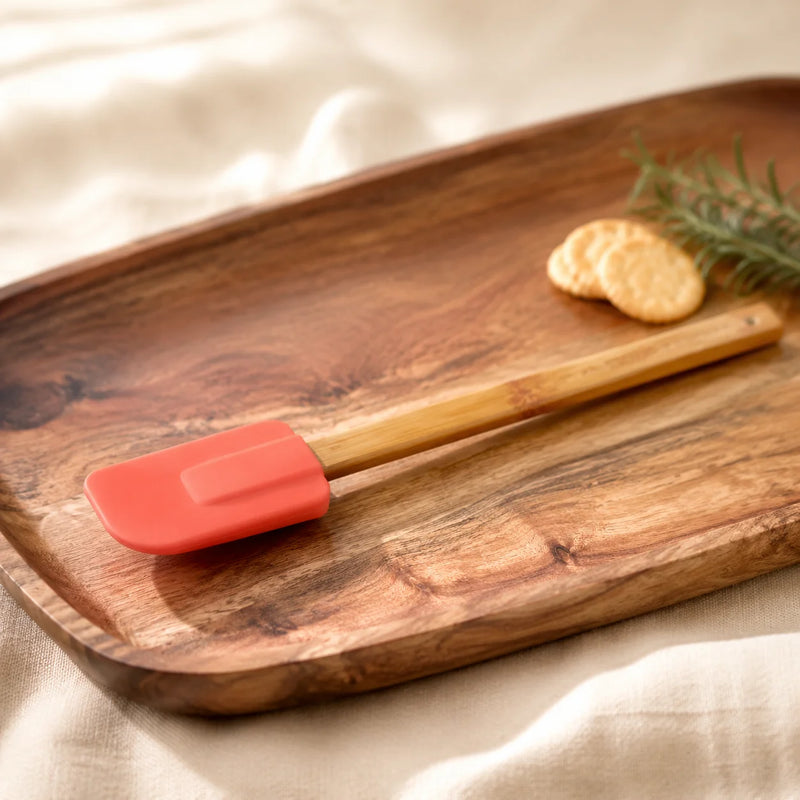 A silicone spatula with a coral-red blade and a light wood handle resting on a rustic wooden serving board, with two round crackers and a sprig of rosemary nearby on a beige fabric surface.