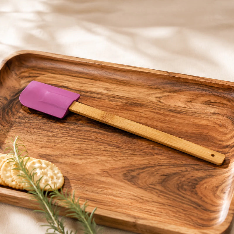 A silicone spatula with a purple silicone head and a light bamboo handle, resting on a rectangular wooden serving tray with crackers and rosemary.