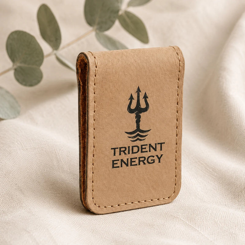A small tan leather card holder or wallet with a black Trident Energy logo and text, standing upright on a light fabric surface with soft eucalyptus leaves in the background.