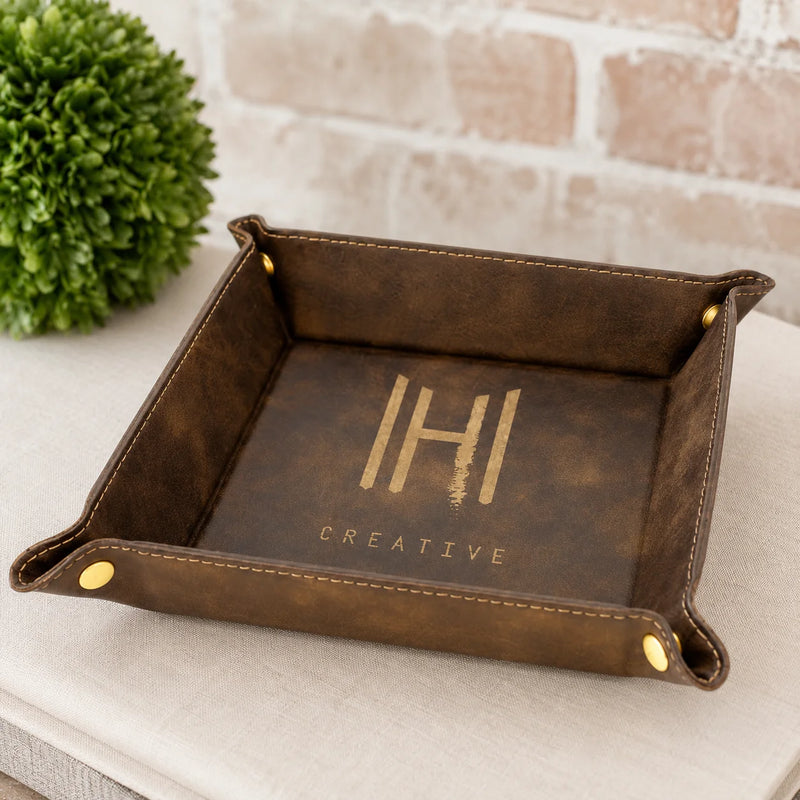 A square brown faux-leather or leather-like tray with rolled corners and gold-tone metal snap accents, placed on a light fabric surface. The inside of the tray is printed with a large stylized 'HI' and the word 'CREATIVE'.