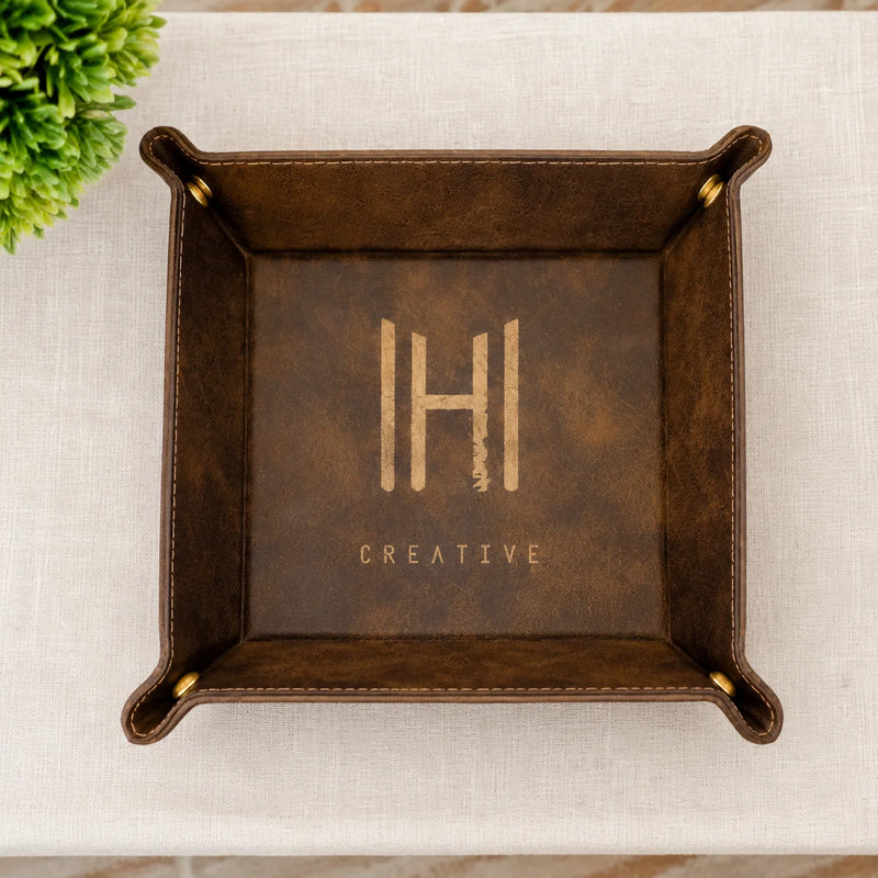 A square dark brown faux-leather tray with rolled corners and brass grommets, featuring a gold foil monogram 'IH' and the word 'CREATIVE' on the bottom, shown from a top-down view on a light beige textured surface with a small green plant in the corner.