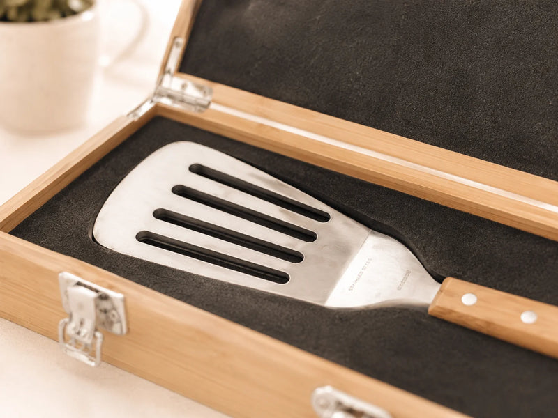 A stainless steel slotted spatula with a light wood handle resting inside an open bamboo presentation box with black foam lining.