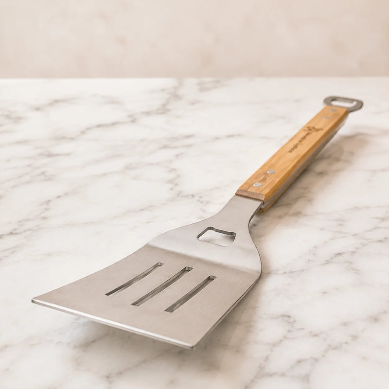 A stainless steel slotted spatula with a wooden handle and bottle opener cutouts, resting on a white marble surface.