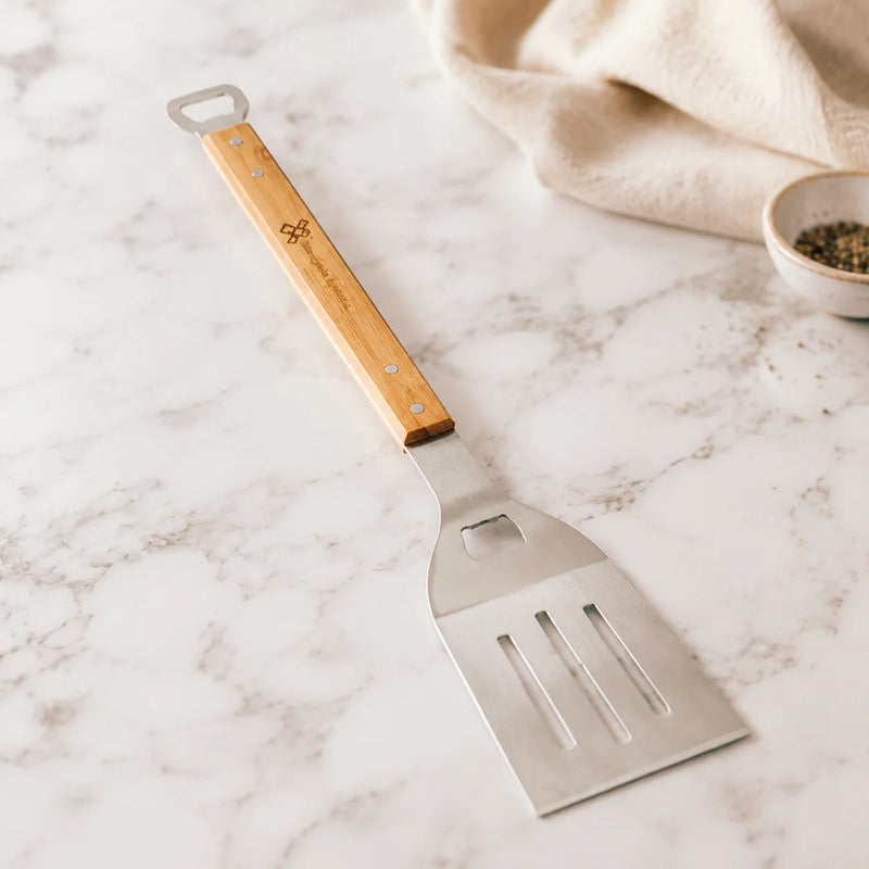 A stainless steel spatula with a light wood handle lies diagonally on a white marble surface. The handle is engraved with the brand name 'BONSAI' and a small logo.
