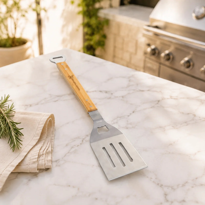 A stainless steel spatula with a light wood handle rests on a white marble outdoor kitchen countertop, with a folded beige towel and rosemary nearby and a grill visible in the background.