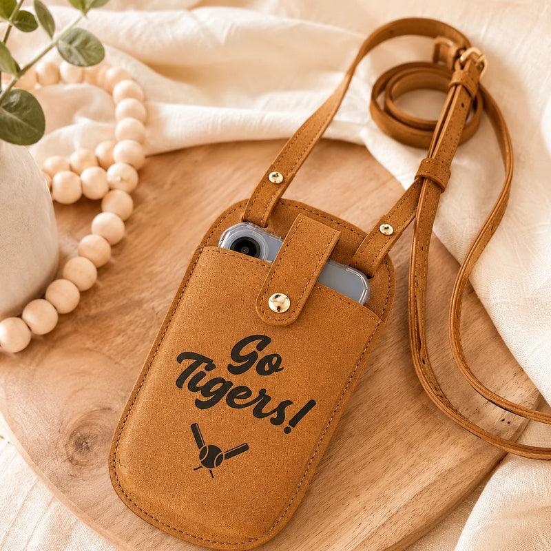A tan faux-leather crossbody phone pouch with a strap, featuring the text 'Go Tigers!' and a crossed bats graphic in black. The pouch is shown holding a silver smartphone on a wooden board with neutral-toned fabric and decorative beads in the background.