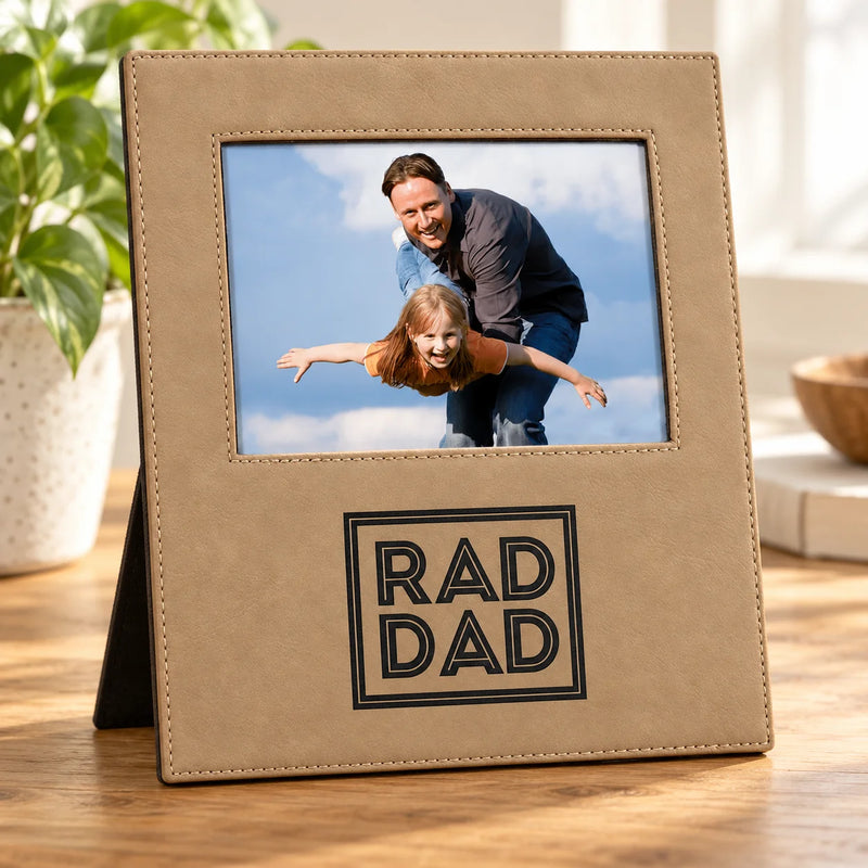 A tan faux-leather photo frame with a stand, featuring a custom photo of a father and daughter and the text 'RAD DAD' in black. The frame is displayed on a wooden table with a potted plant in the background.