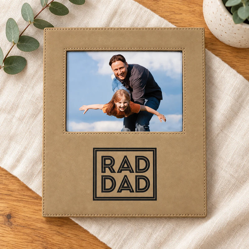 A tan faux-leather picture frame with a custom photo of a father and daughter and the engraved text 'RAD DAD' on the front, styled on a cream linen cloth over a wooden table.