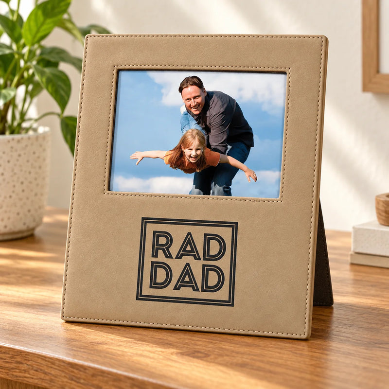 A tan faux-leather picture frame with a stitched border and a front panel reading “RAD DAD” in black outlined block letters, standing on a wooden table. The photo insert shows a man and child, and a plant is visible in the background.