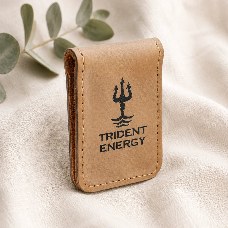 A tan leather card holder wallet with a black trident logo and the text 'TRIDENT ENERGY' printed on the front, resting on a beige fabric background with eucalyptus leaves.