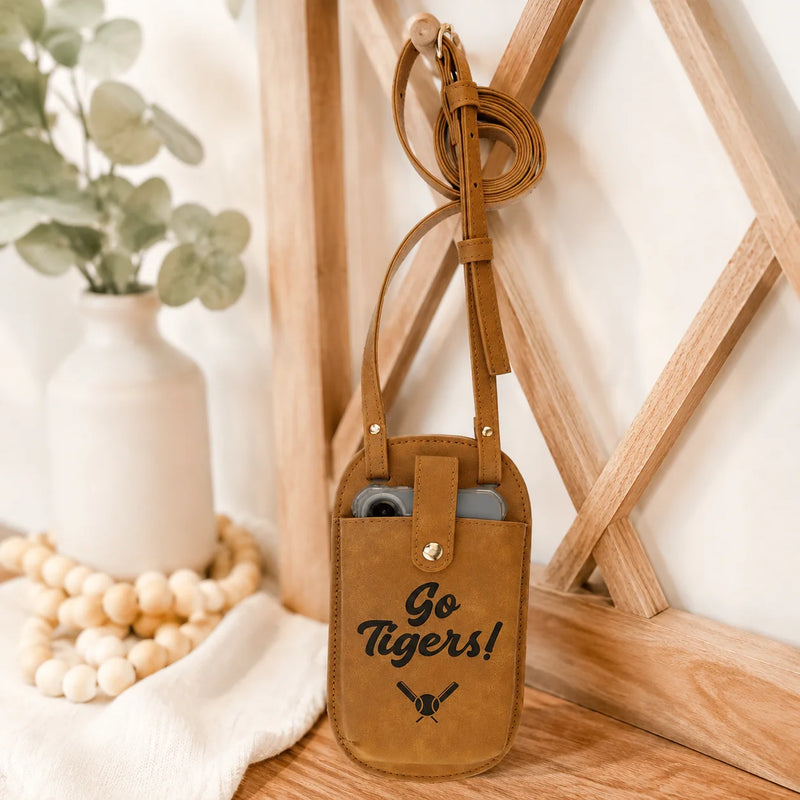 A tan leather crossbody phone pouch with a 'Go Tigers!' graphic, styled on a wooden surface next to a white vase with greenery.