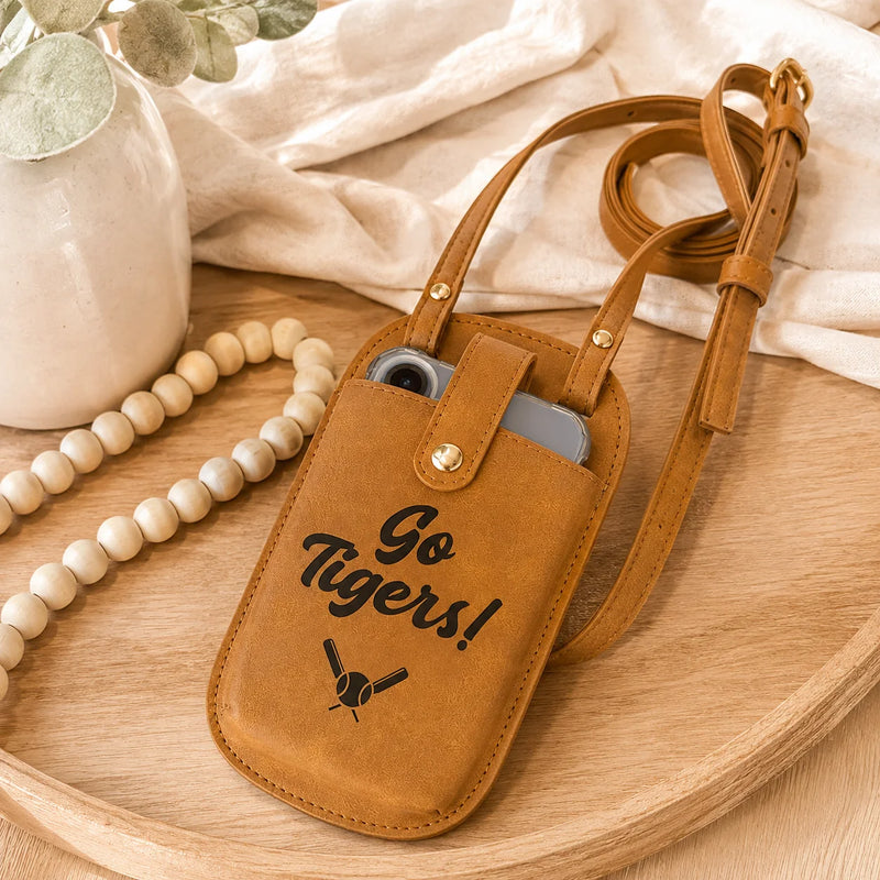 A tan leather crossbody phone pouch with a gold snap closure and adjustable strap, featuring the text 'Go Tigers!' and a tiger graphic. The pouch is styled on a wooden tray with a beaded necklace, a white vase, and draped fabric.