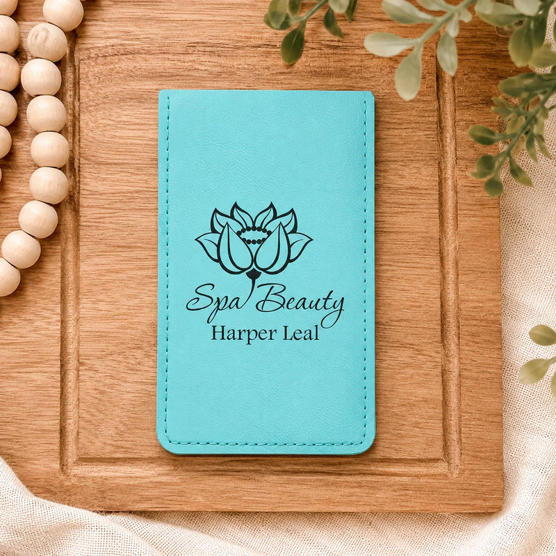 A turquoise blue faux leather passport cover or notebook cover with black printed text reading 'Spa Beauty Harper Leal' and a lotus flower graphic, displayed on a wooden board with decorative beads and greenery.