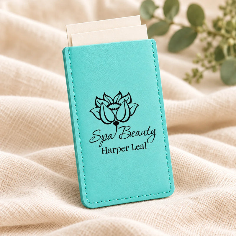 A turquoise faux-leather card holder or wallet with a black lotus logo and the text 'Spa Beauty Harper Leal' printed on the front, standing upright on a beige textured fabric with blurred green leaves in the background.