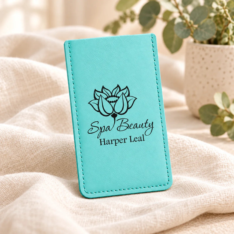 A turquoise faux-leather card holder or wallet with black printed text reading 'Spa Beauty Harper Leal' and a lotus flower graphic, standing on a beige fabric surface with soft-focus greenery in the background.