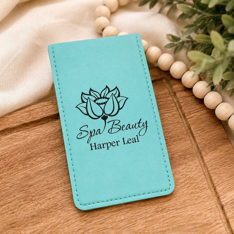 A turquoise faux-leather card holder or wallet with black printed text reading 'Spa Beauty Harper Leal' and a lotus flower graphic, styled on a wooden surface with a beige cloth, wooden beads, and greenery.